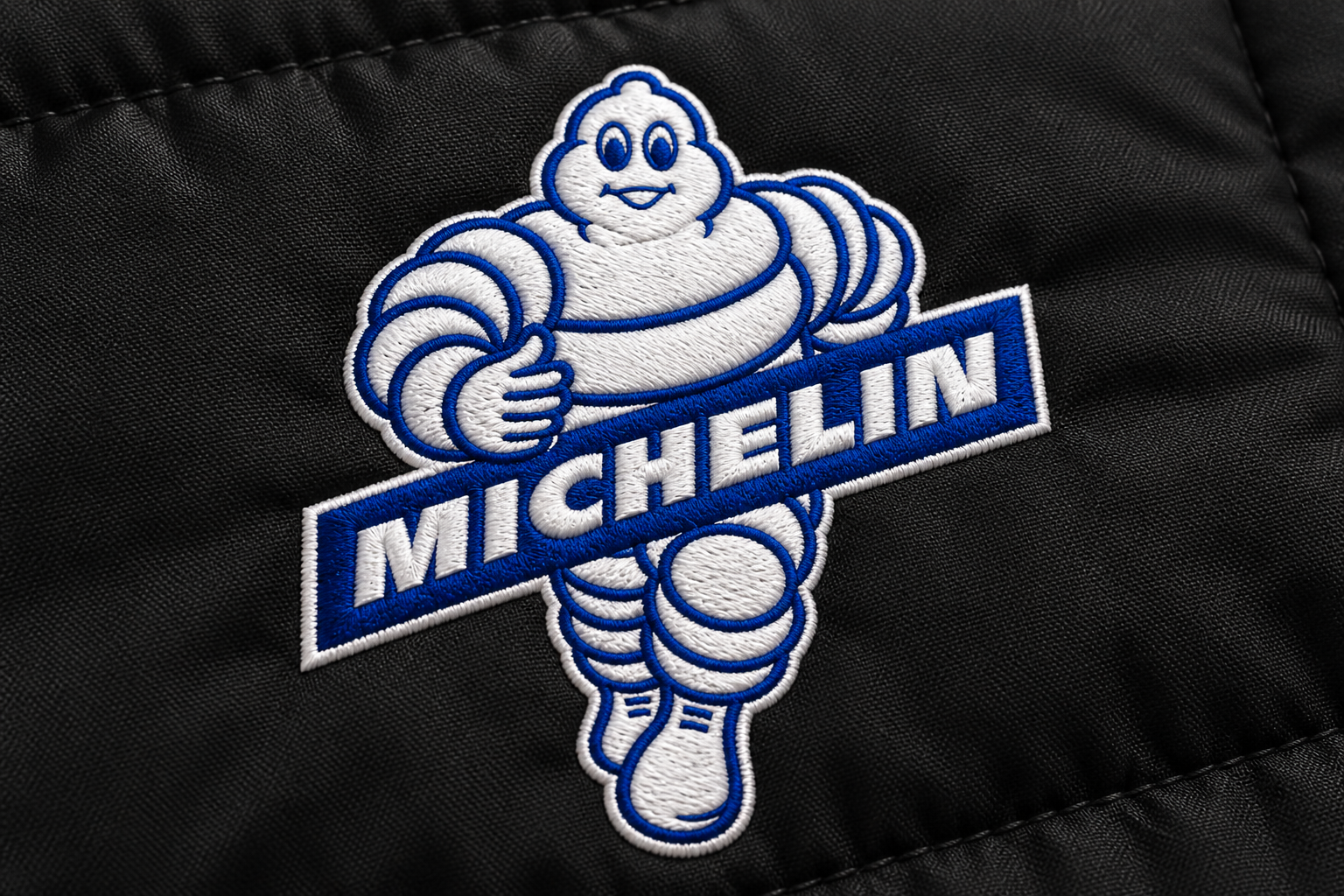Gros plan d’une simulation de broderie Michelin en fils blancs et bleus sur un textile noir, avec le relief des points visible